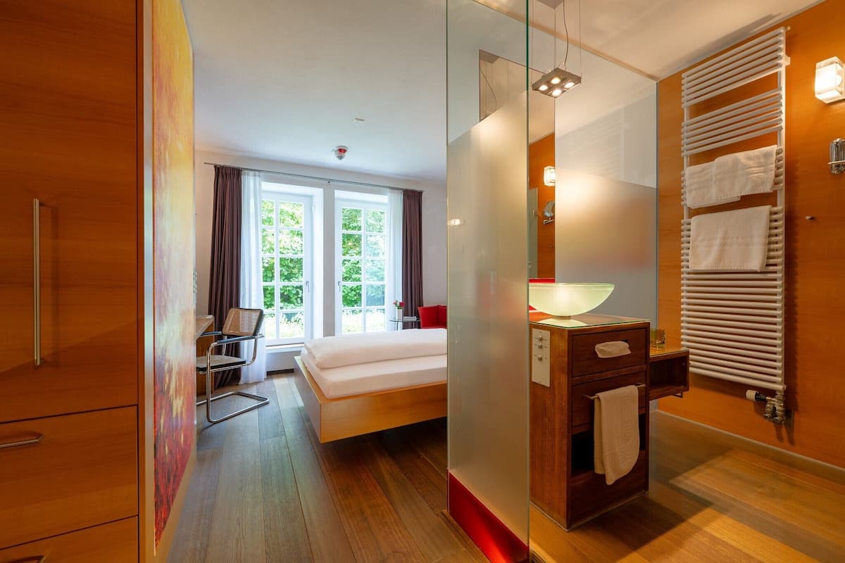 Een moderne hotelkamer met een bed, zithoek, badkamer met handdoekhouder en uitzicht op groen.