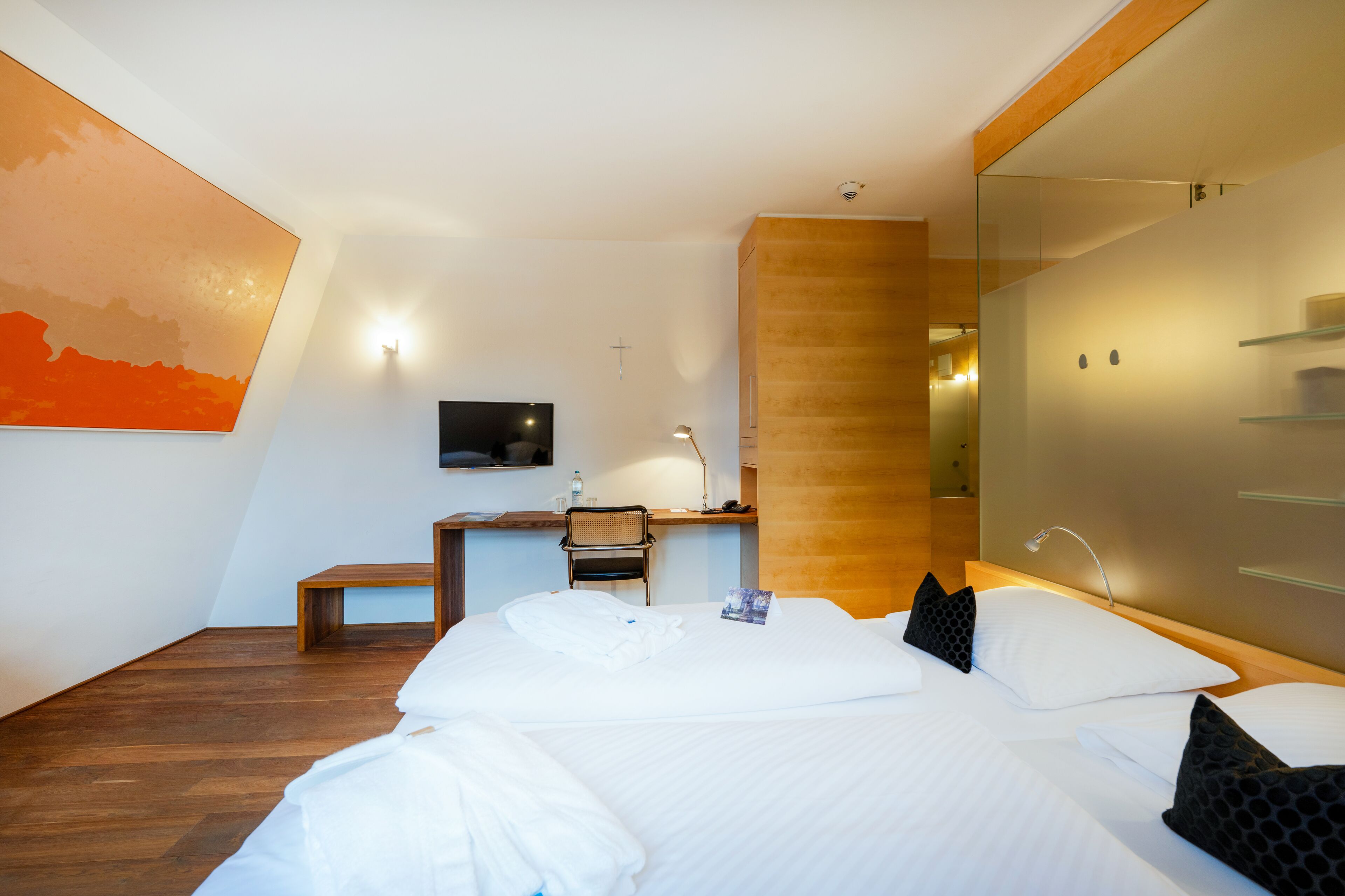A modern hotel room with a large bed, a desk with a chair, a television, and a glass partition.