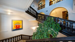Een statige trap met een zwarte balustrade, een grote groene plant en een lijstwerk met een kunstwerk aan de muur.