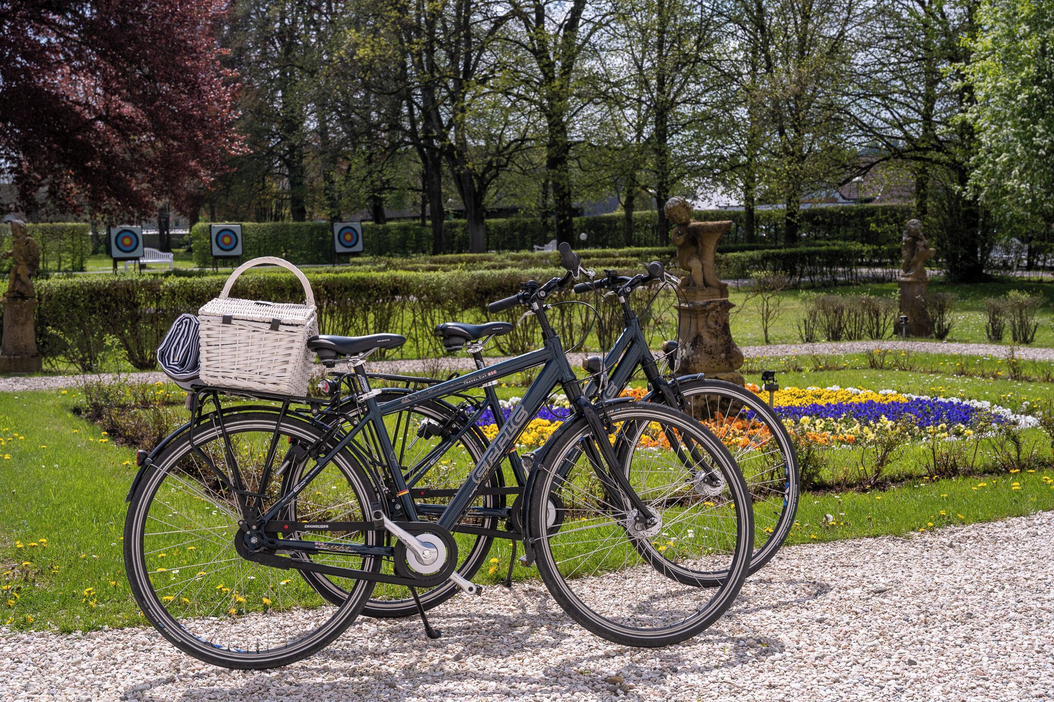 A bicycle with a basket and a seat, parked in a garden with flower beds and a hedge.
