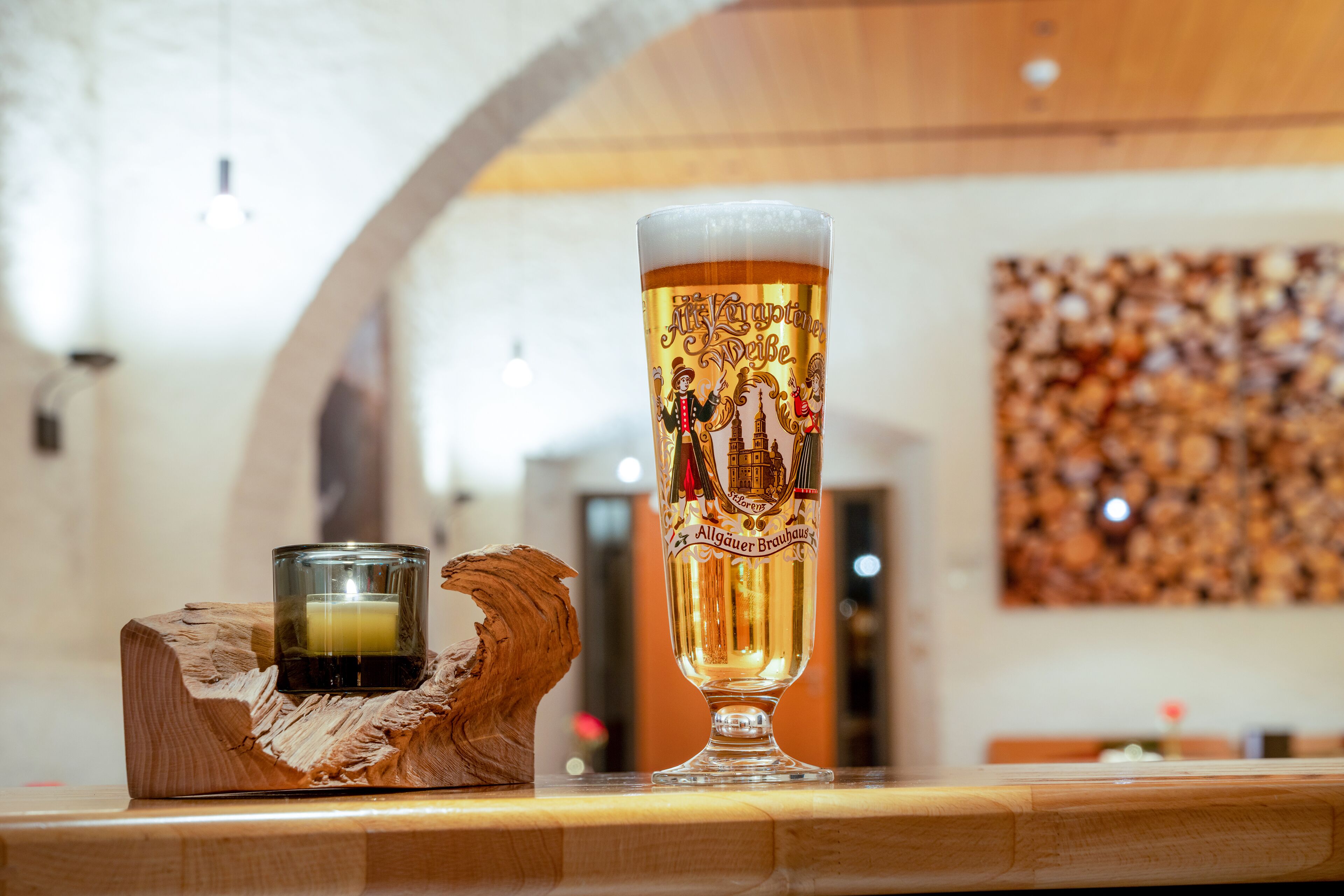 Een hoog glas bier met een logo, een kaars en een houten beeldje op een tafel.