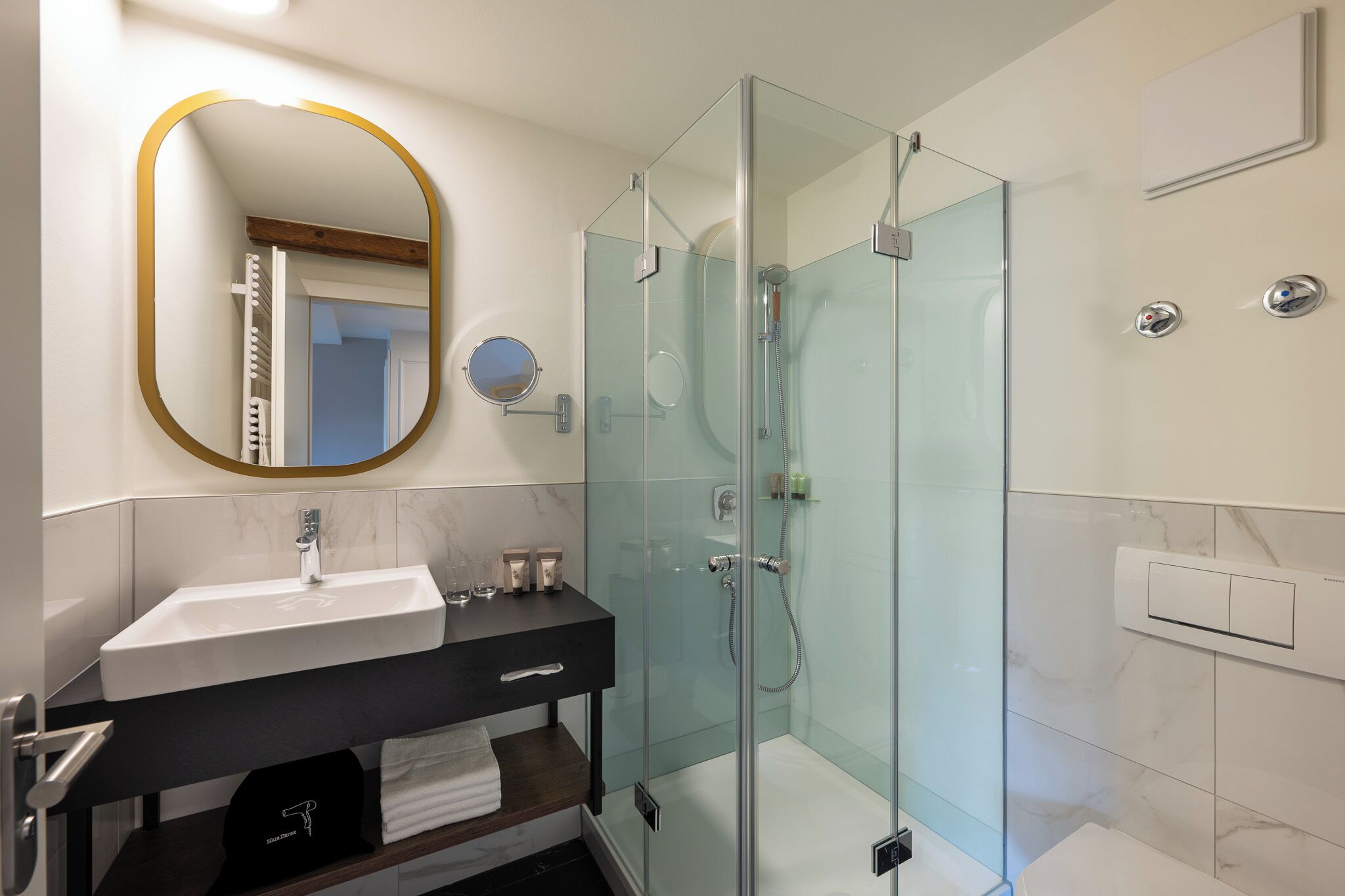 Un baño moderno con ducha envuelta en vidrio, un espejo ovalado grande y un mueble de tocador negro con encimera de mármol.