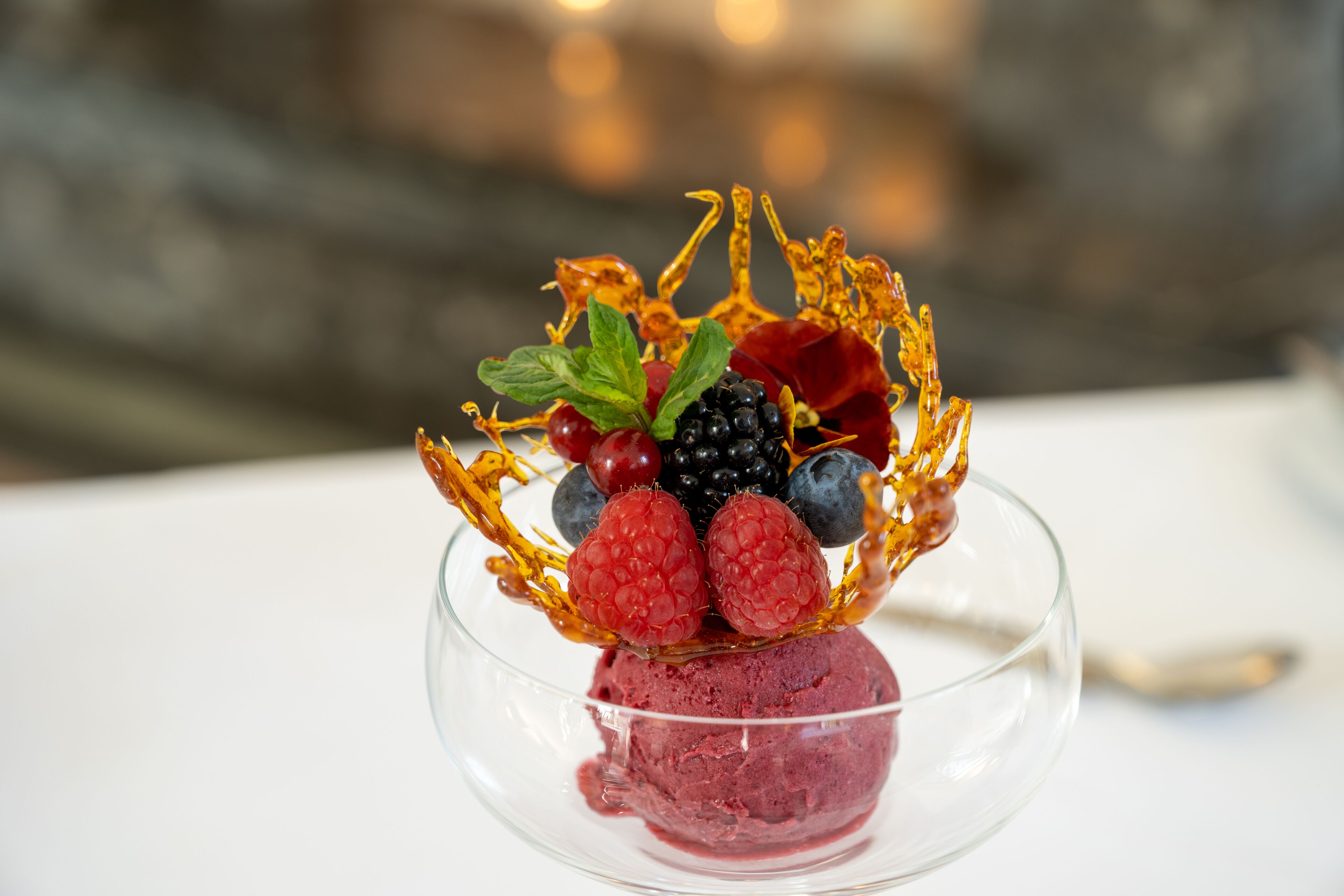 Un postre con bayas y una hoja dorada como decoración en un bol de cristal transparente.