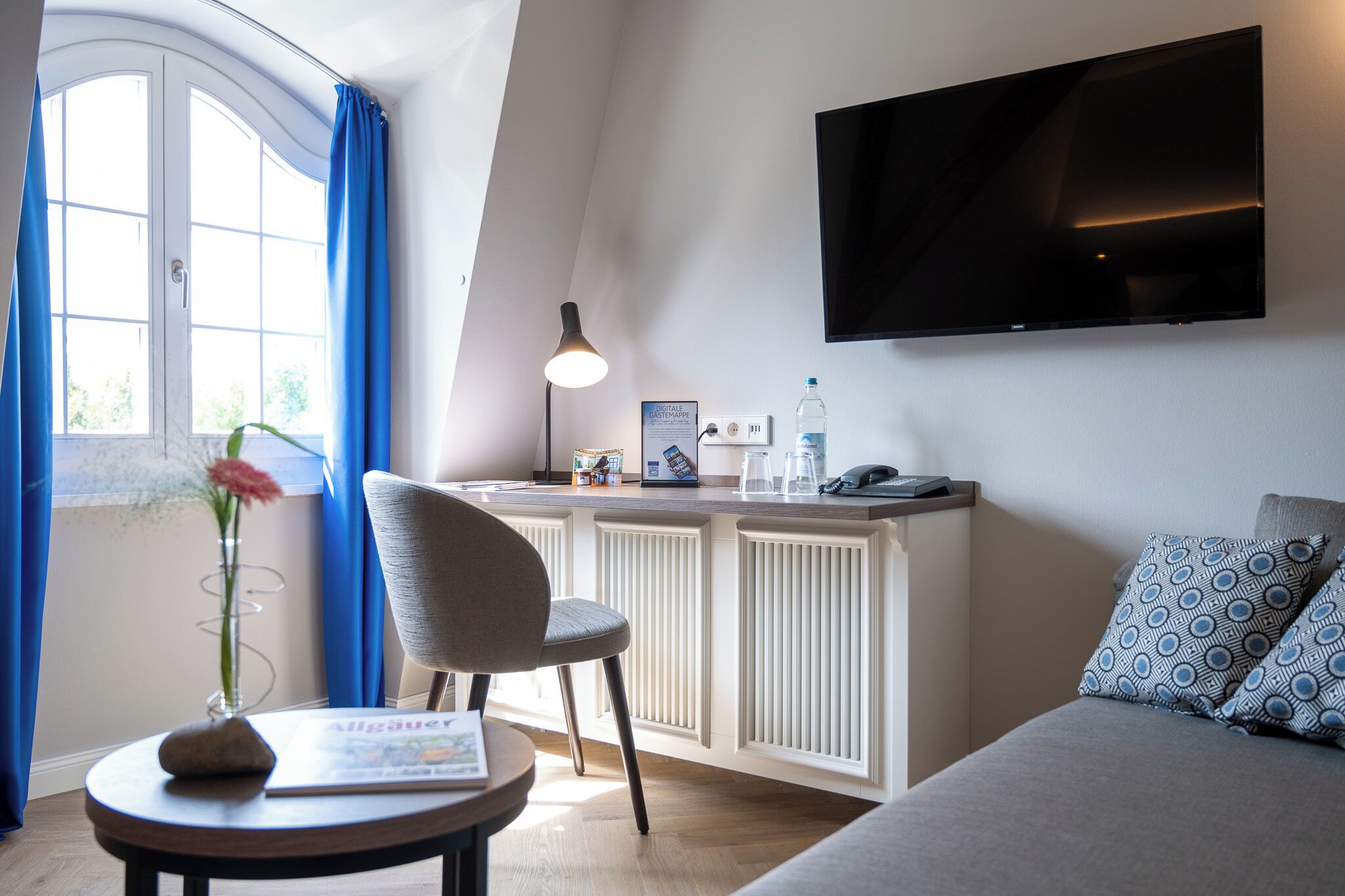Une chambre moderne dotée d’un téléviseur à écran plat fixé au mur, d’un petit coin cuisine, d’un espace salon avec un canapé et un fauteuil, et d’une fenêtre avec des rideaux.