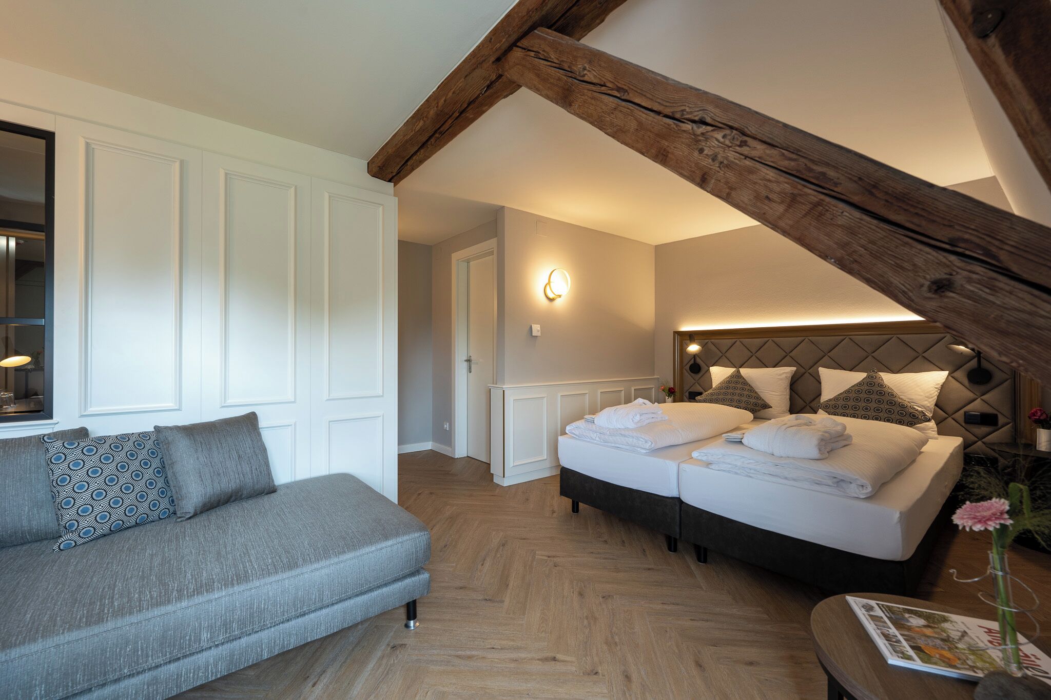 Ein Schlafzimmer mit Holzbalkendecke, einer Couch, einem Bett mit Kissen und einem Nachttisch mit einer Zeitschrift.
