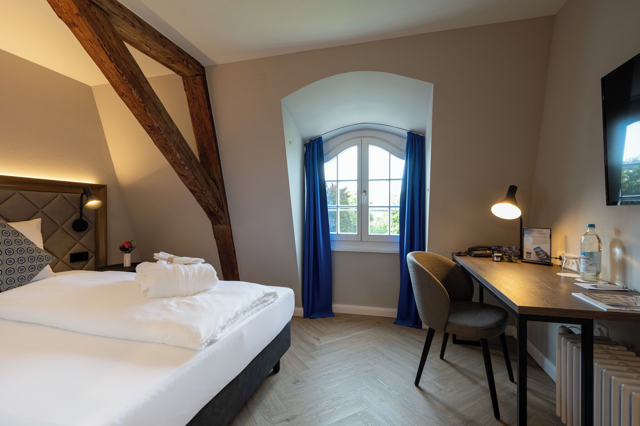 A hotel room with a bed, a desk, a chair, a window with curtains, and a mounted TV.