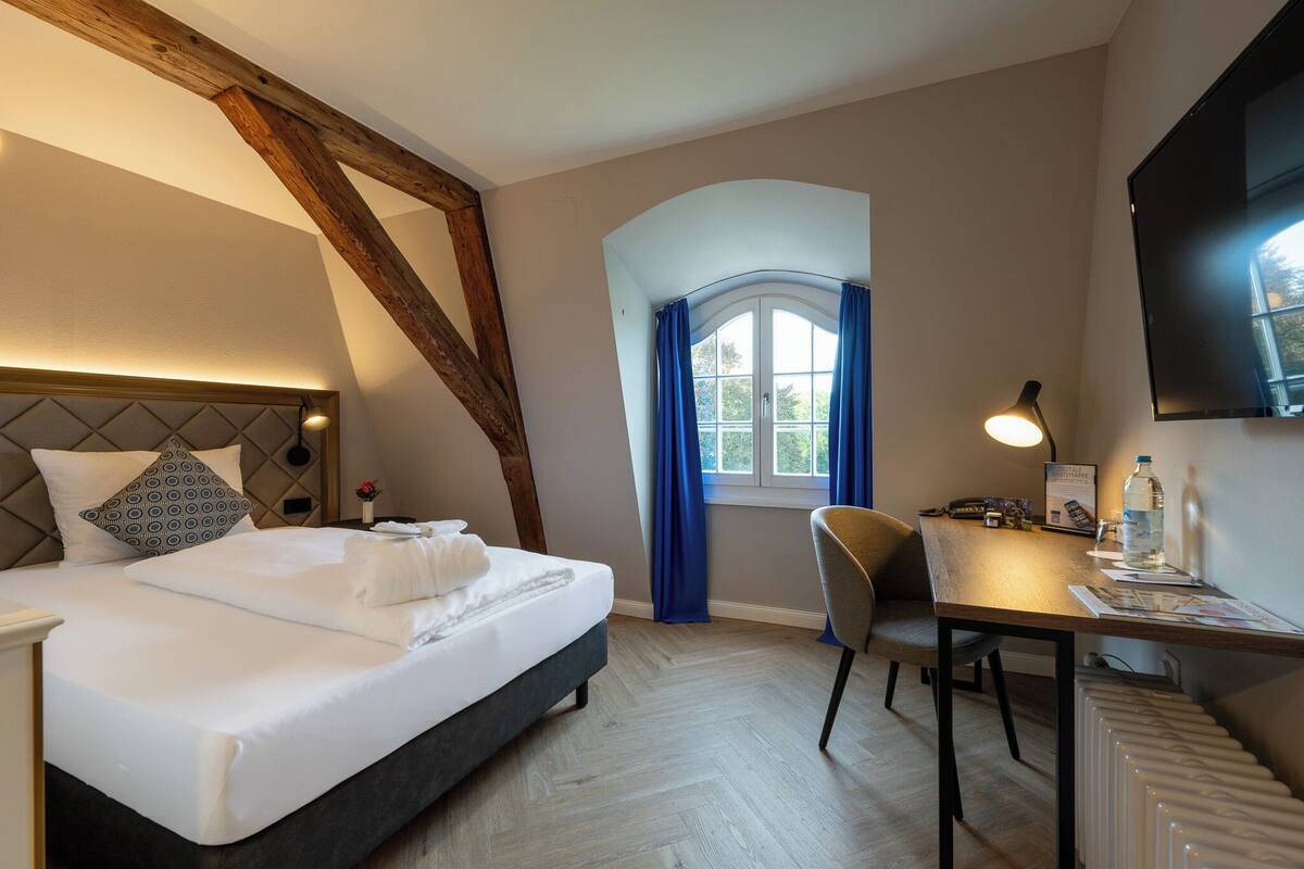 Een hotelkamer met een bed, een bureau, een stoel, een raam met gordijnen, een televisie en een radiator.