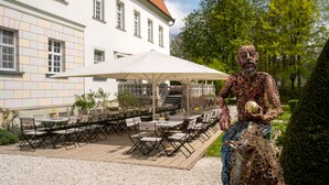 Een terras met tafels, stoelen, een grote parasol en een standbeeld.
