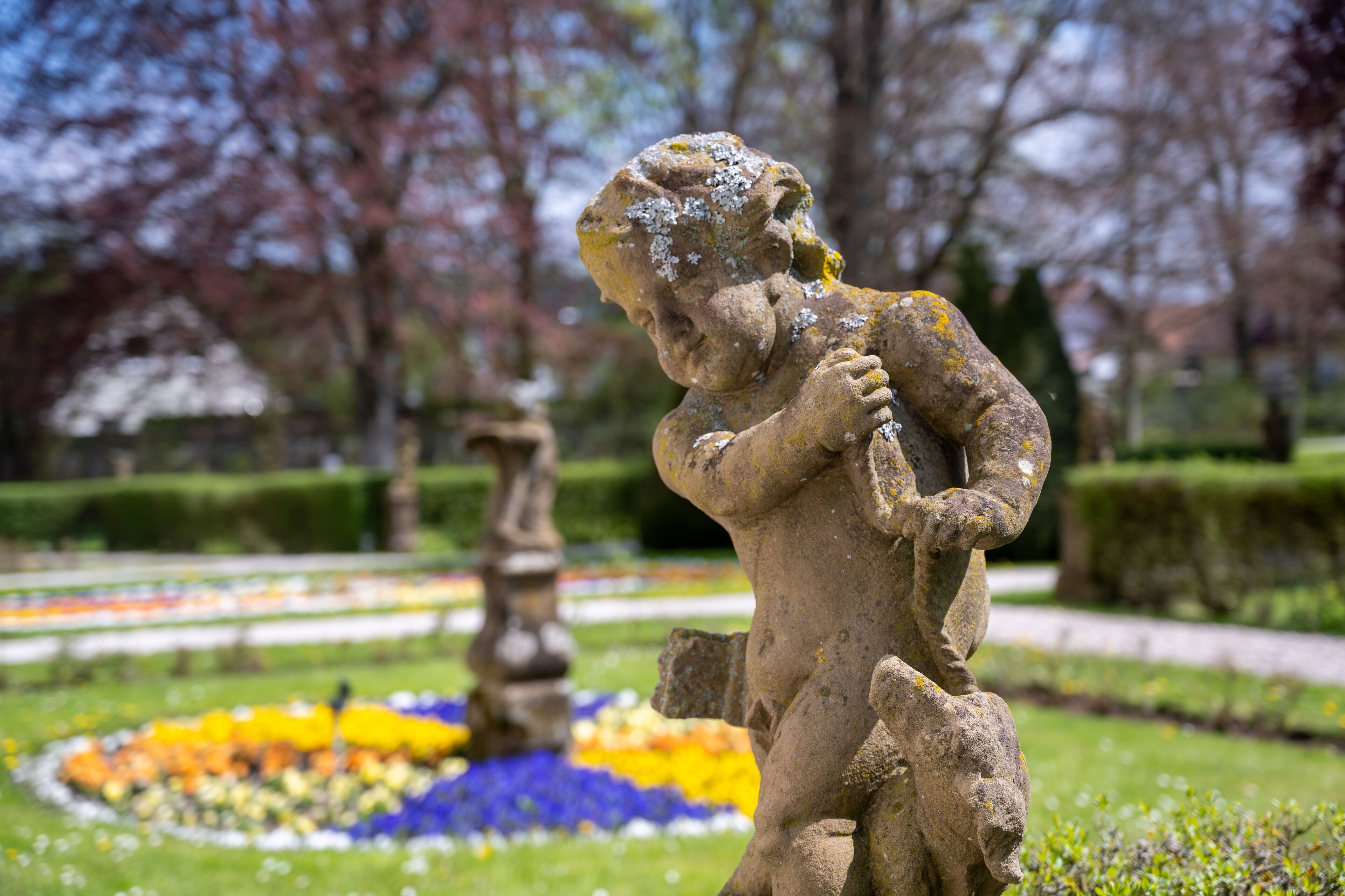Une statue en pierre représentant un enfant, placée dans un jardin, avec un parterre de fleurs circulaire en arrière-plan.