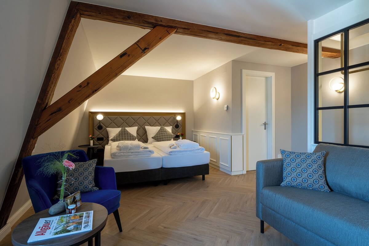 Een slaapkamer met een bed, een bank, een kleine tafel met een tijdschrift en een decoratieve plant.