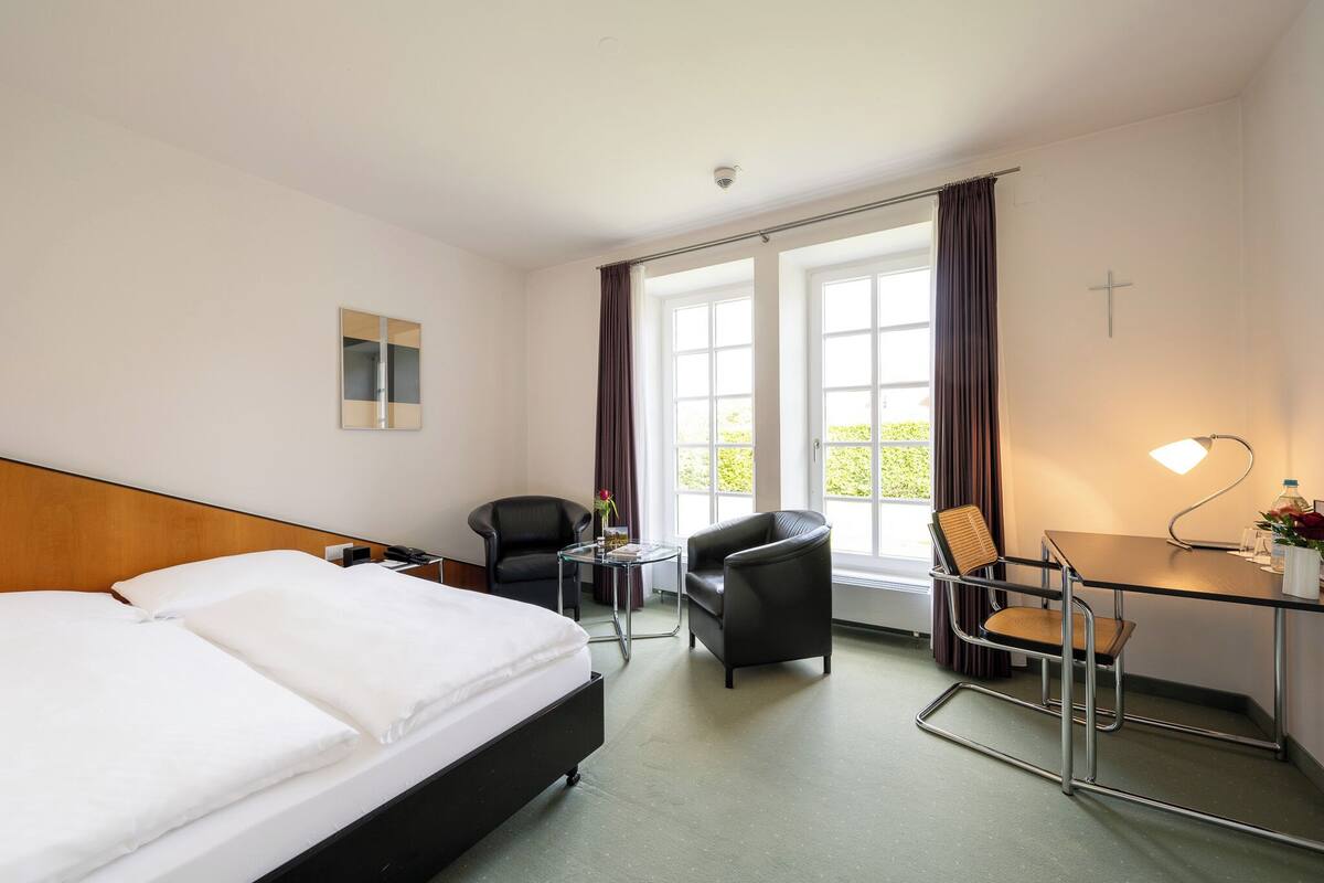 Een hotelkamer met een bed, twee stoelen, een bureau en een groot raam met gordijnen.