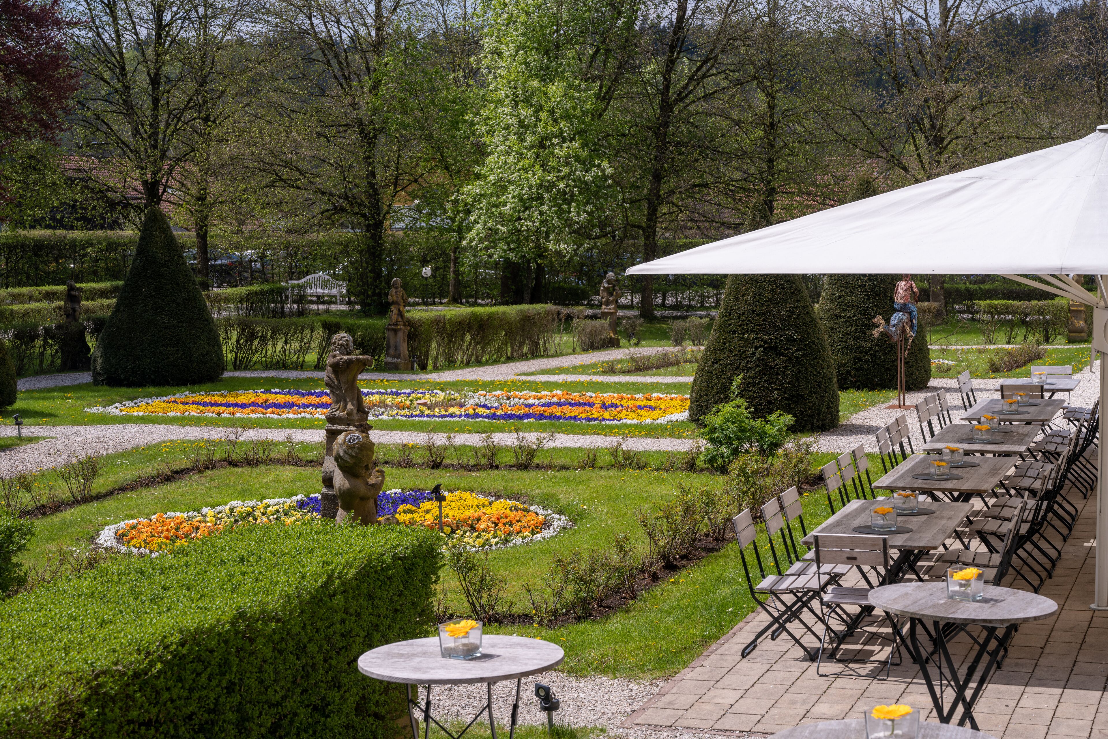 An outdoor seating area with tables and chairs, surrounded by manicured gardens and a statue.