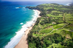 Vue aérienne d’une zone côtière avec une plage, des champs verdoyants et quelques bâtiments dispersés.
