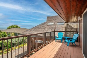 Un balcón con terraza de madera, dos sillas Adirondack azules y vista al océano y edificios cercanos.