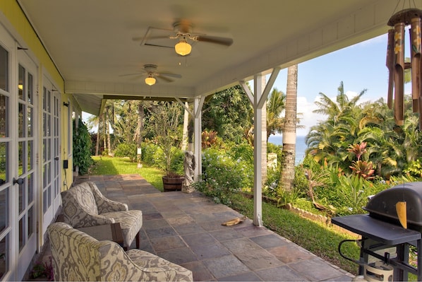 Une terrasse couverte, aménagée avec du mobilier d’extérieur, un barbecue et offrant une vue sur un jardin luxuriant et l’océan.
