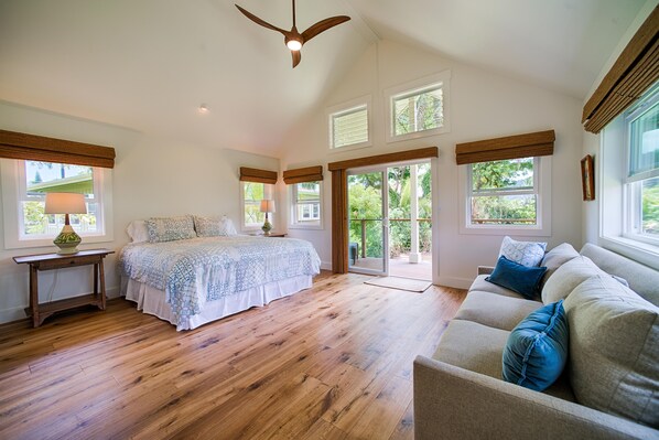 A spacious bedroom with a large bed, a wooden floor, a ceiling fan, and a view through the glass doors.