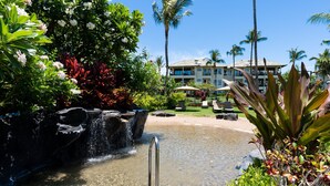 Zona de piscina en un resort con cascada, rodeada de exuberante vegetación tropical y palmeras.