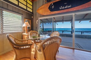 A dining area with wicker chairs and a glass table, overlooking a beach with a wooden boat sign.