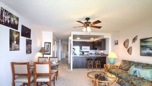 A living room with a sofa, a coffee table, and a dining area with chairs. There is a kitchen in the background with a microwave and a ceiling fan. - Maalaea Kai 316 1 Bedroom Condo by RedAwning (Wailuku)