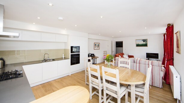 A modern kitchen with white cabinets, a dining area with a wooden table and chairs, and a living area with a sofa and red curtains. - Up-market one Bedroom Apartment Just Minutes From the River Thames. Broughton rd (London)