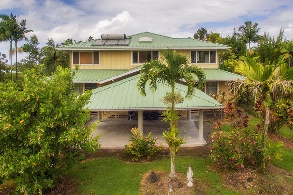 A two-story house with a green roof, surrounded by lush greenery and palm trees. - Maluhia Hale 3 Bedroom Home by RedAwning (Keaau)