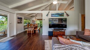A spacious living area with a large TV, wooden furniture, and a balcony with outdoor seating. - Hale Naninoa 5 Bedroom Home by RedAwning (Hanalei)