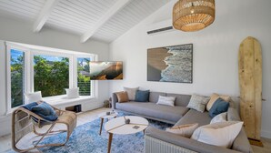 Ein modernes Wohnzimmer mit einem großen Fenster, einem grauen Sofa, einem Korbstuhl und einem Holzsurfbrett.