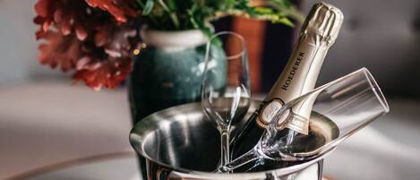 Un balde metálico con una botella de champán y copas, un cuenco de fruta y un jarrón con flores sobre una mesa.