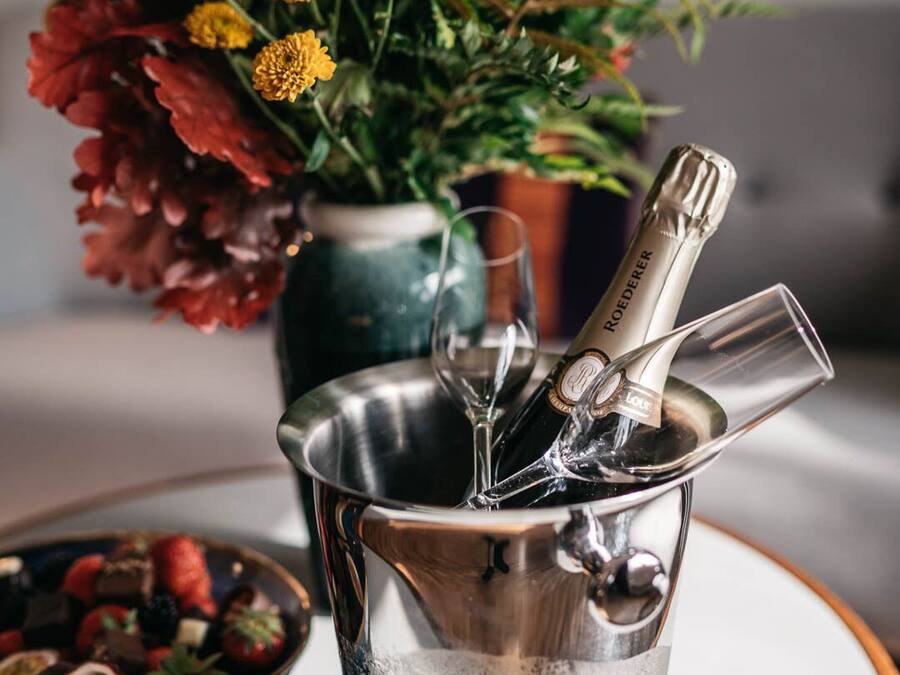Un balde metálico con una botella de champán y copas, un cuenco de fruta y un jarrón con flores sobre una mesa.