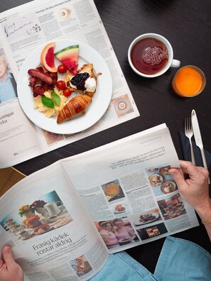 A person reading a magazine with a plate of food, a cup of tea, and a glass of juice on a table. - Rönneberga (Lidingo)