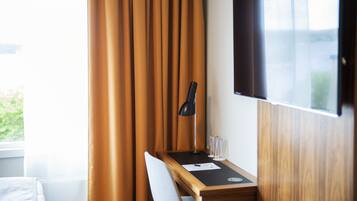 A hotel room with a wooden desk, a black desk lamp, a flat-screen TV mounted on the wall, and a window with curtains.