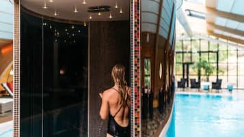 Une femme en maillot de bain noir, dans un espace de douche clos par des parois de verre, avec une piscine visible à l’arrière-plan.