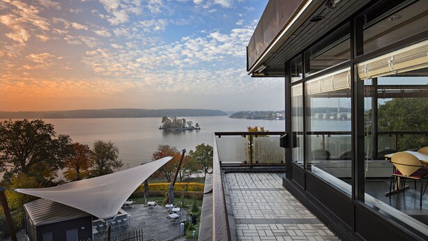 A balcony with a view of a lake and distant islands, a covered outdoor seating area, and a modern building facade. - Rönneberga (Lidingo)