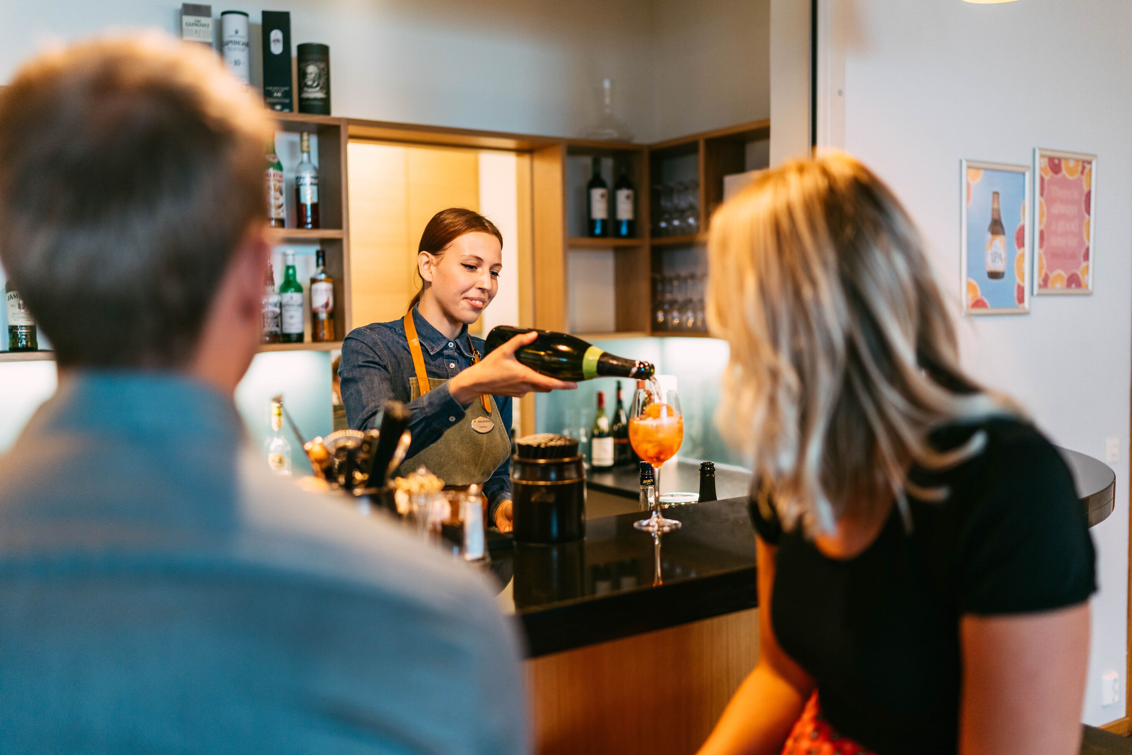 Un coin bar où un barman verse une boisson dans un verre. On peut voir des étagères remplies de bouteilles et de verres derrière le barman.