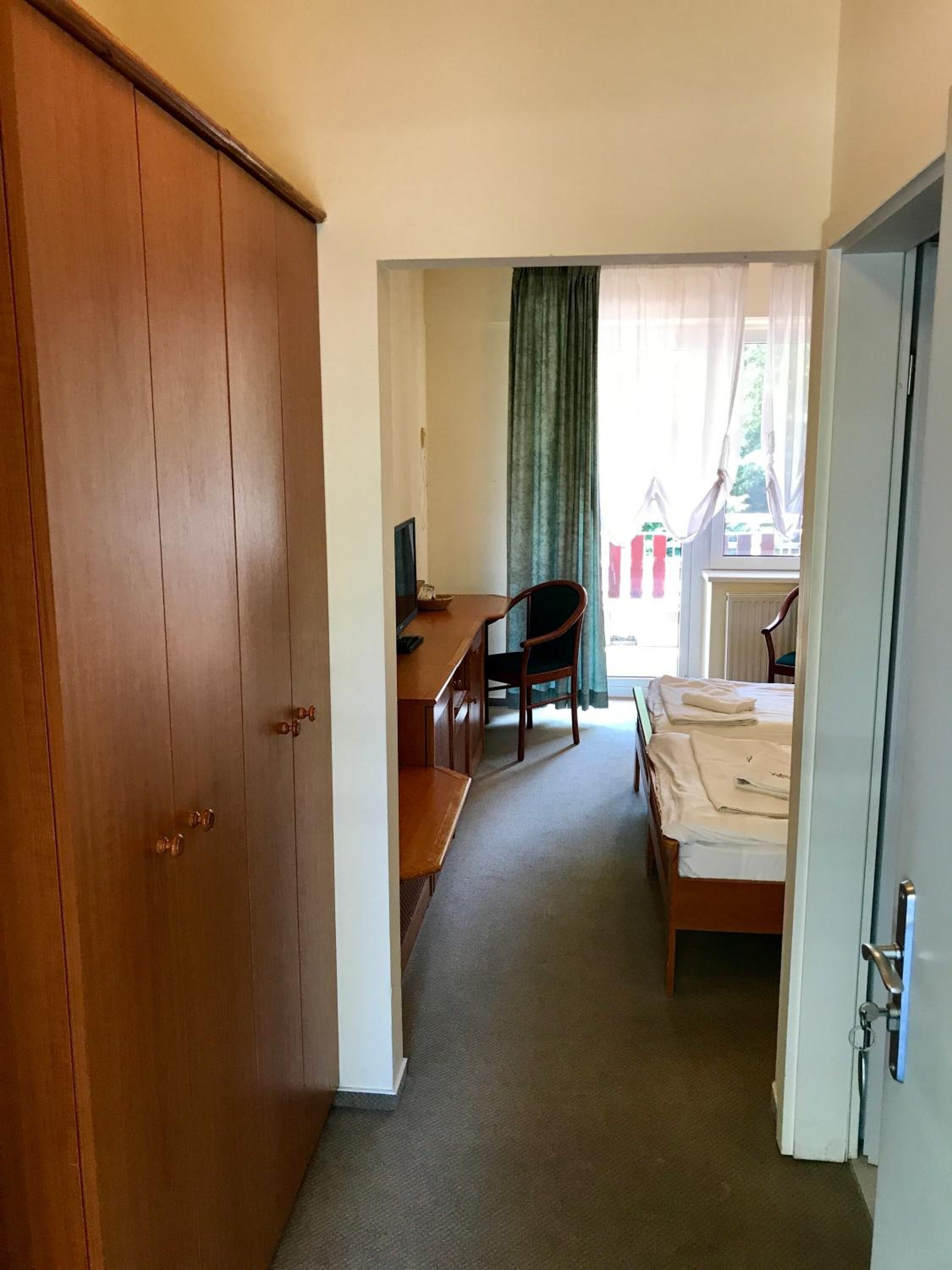 A hotel room with a wooden wardrobe, a desk with a chair, a bed with white linens, and a window with curtains.
