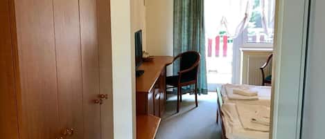 A hotel room with a wooden wardrobe, a desk with a chair, a bed with white linens, and a window with curtains.