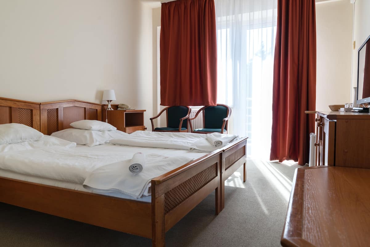 Habitación de hotel con cama de madera, dos sillas, un escritorio y un ventanal con cortinas.