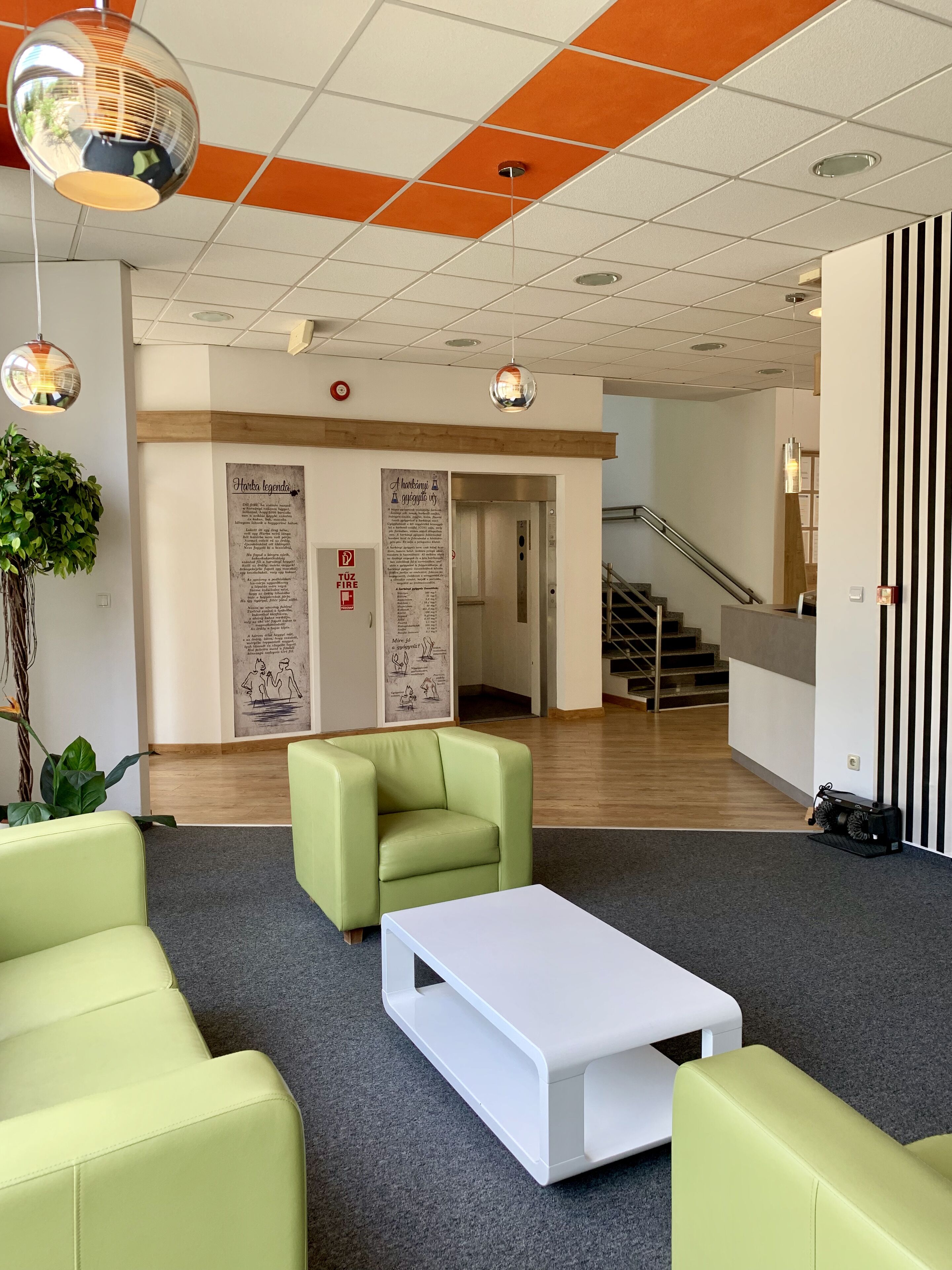 A lobby with green sofas, a white coffee table, and a plant. There are signs on the wall and a staircase in the background.