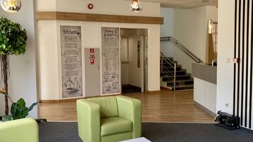 A lobby with green sofas, a white coffee table, and a plant. There are signs on the wall and a staircase in the background.