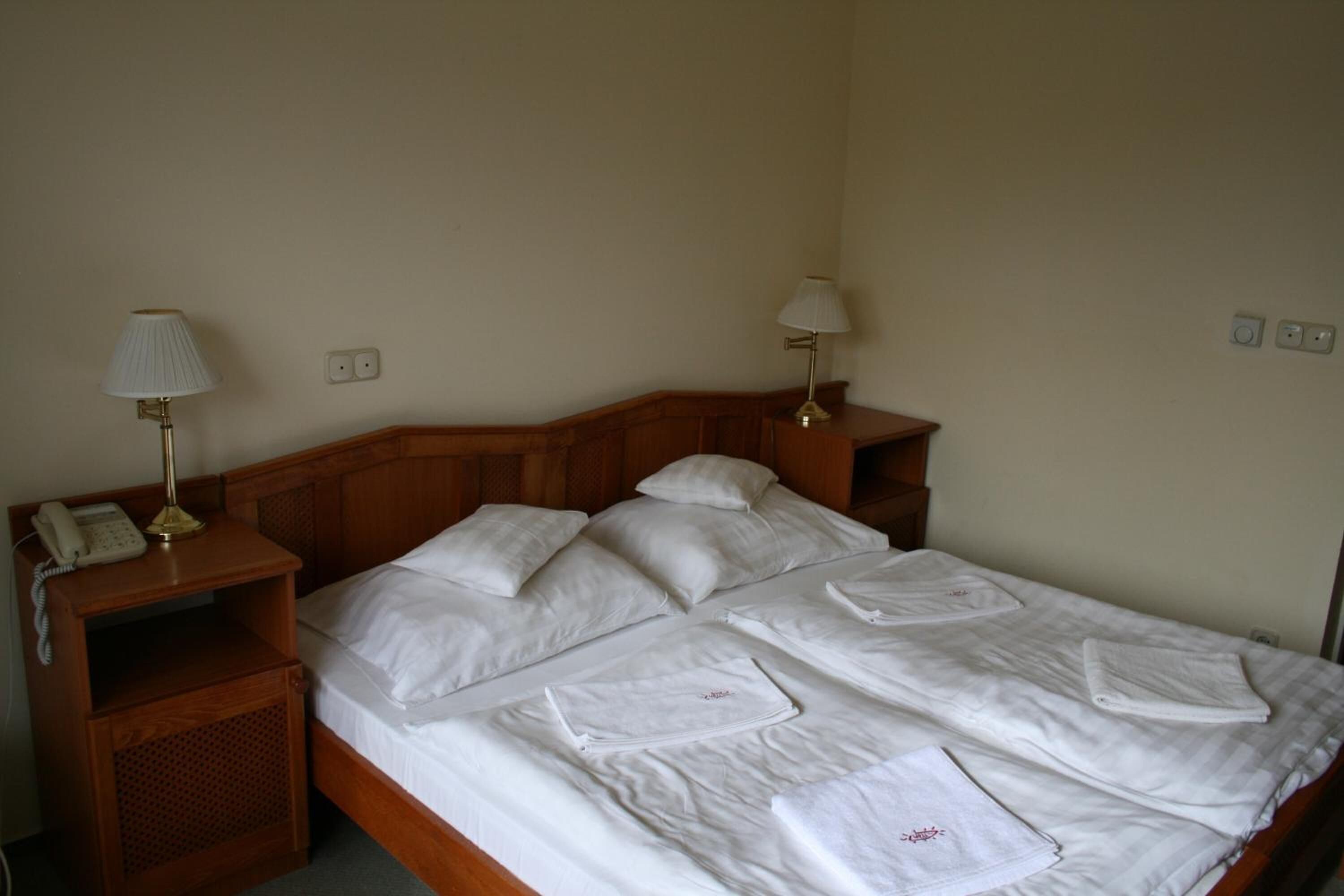 A double bed with white linens, two bedside lamps, a wooden nightstand with a telephone, and a small shelf.