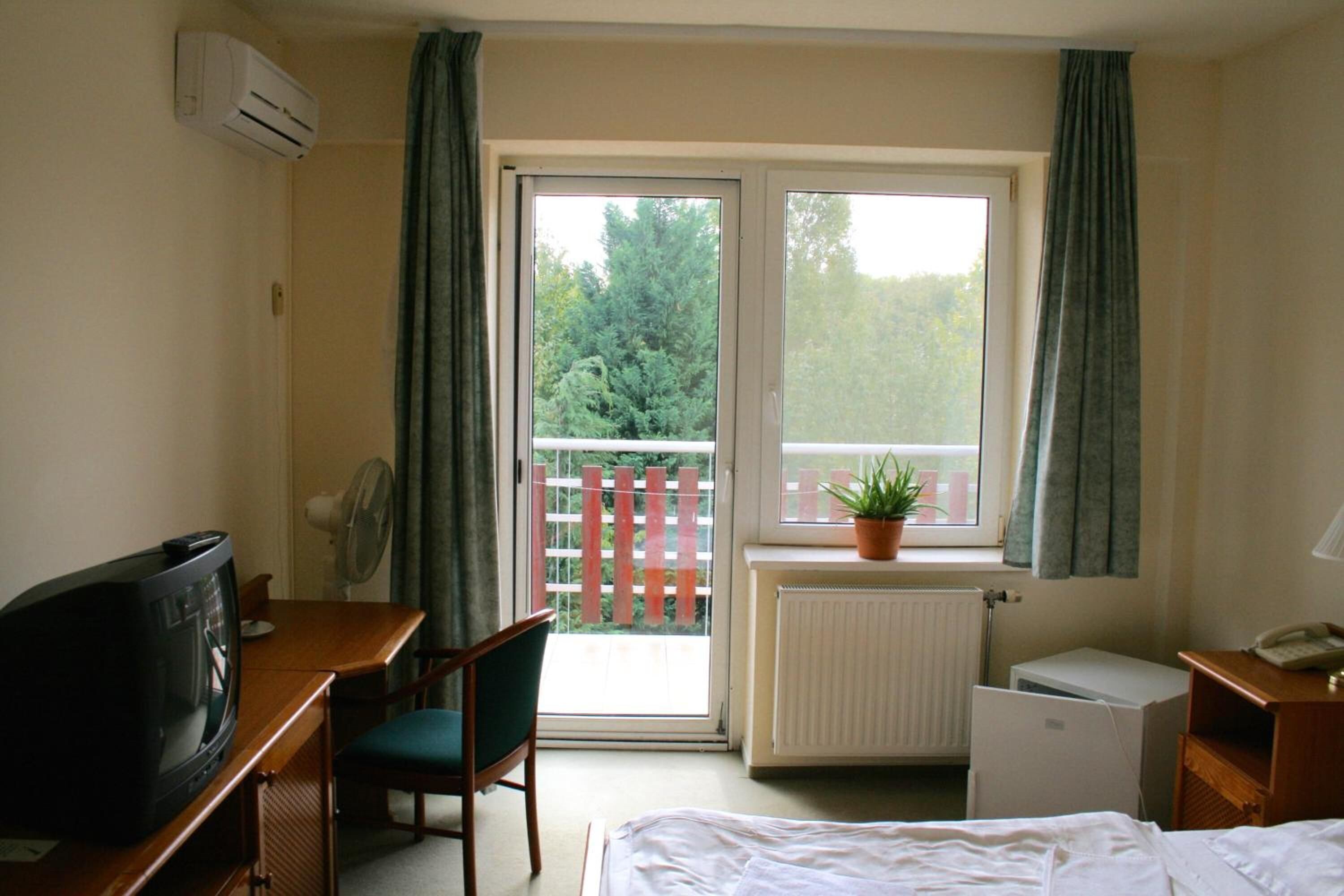 Habitación de hotel con una cama, un escritorio con televisor, una silla, un radiador y una ventana con vistas a árboles y una valla.