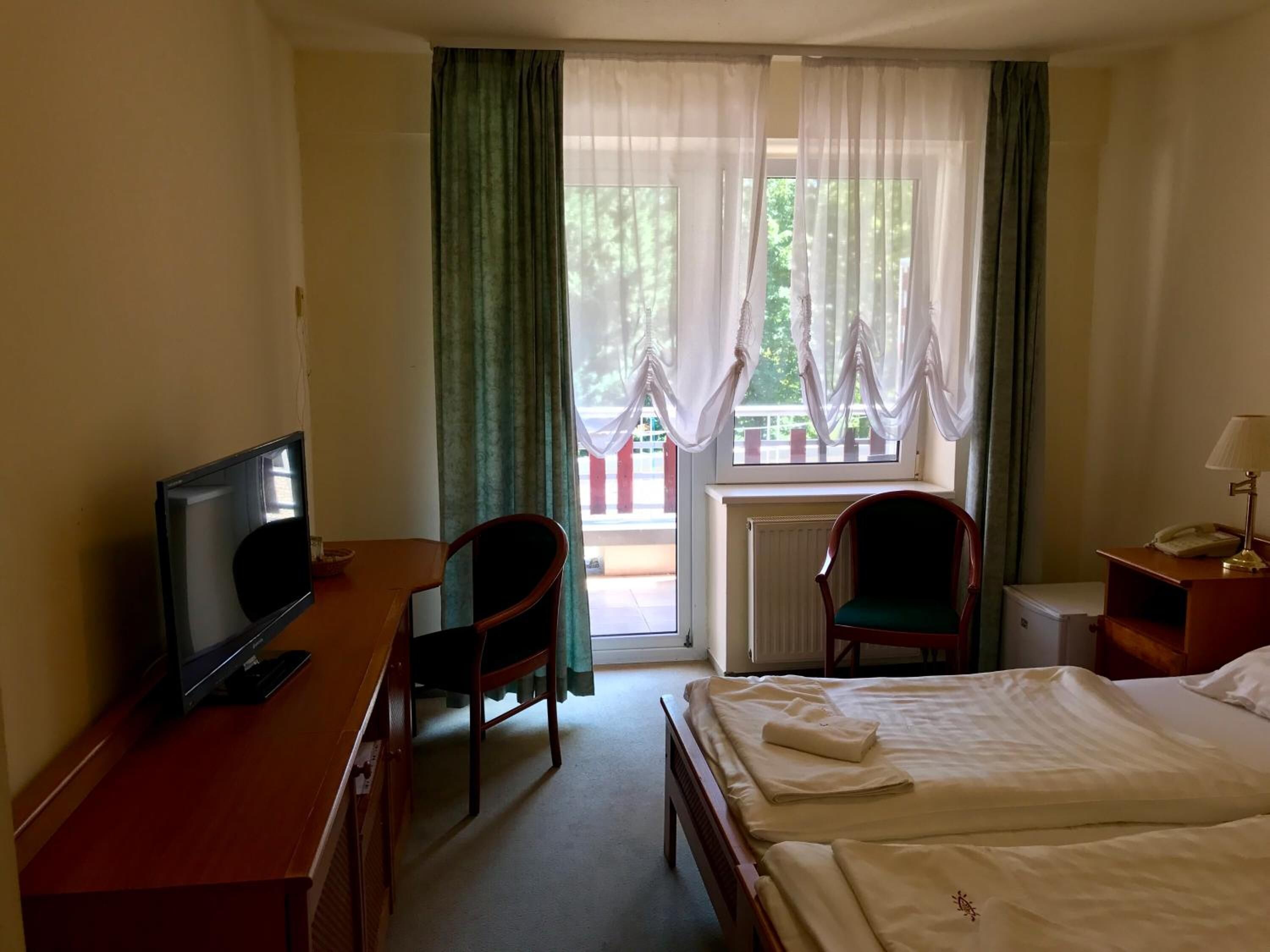 Habitació d'hotel amb un llit, una escriptori amb televisió, dues cadires, una petita taula de nit amb una làmpada i una finestra amb cortines.