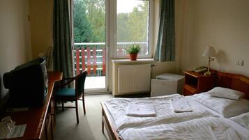A hotel room with a bed, a desk with a television, a chair, a small table with a lamp, and a window with a view of trees.