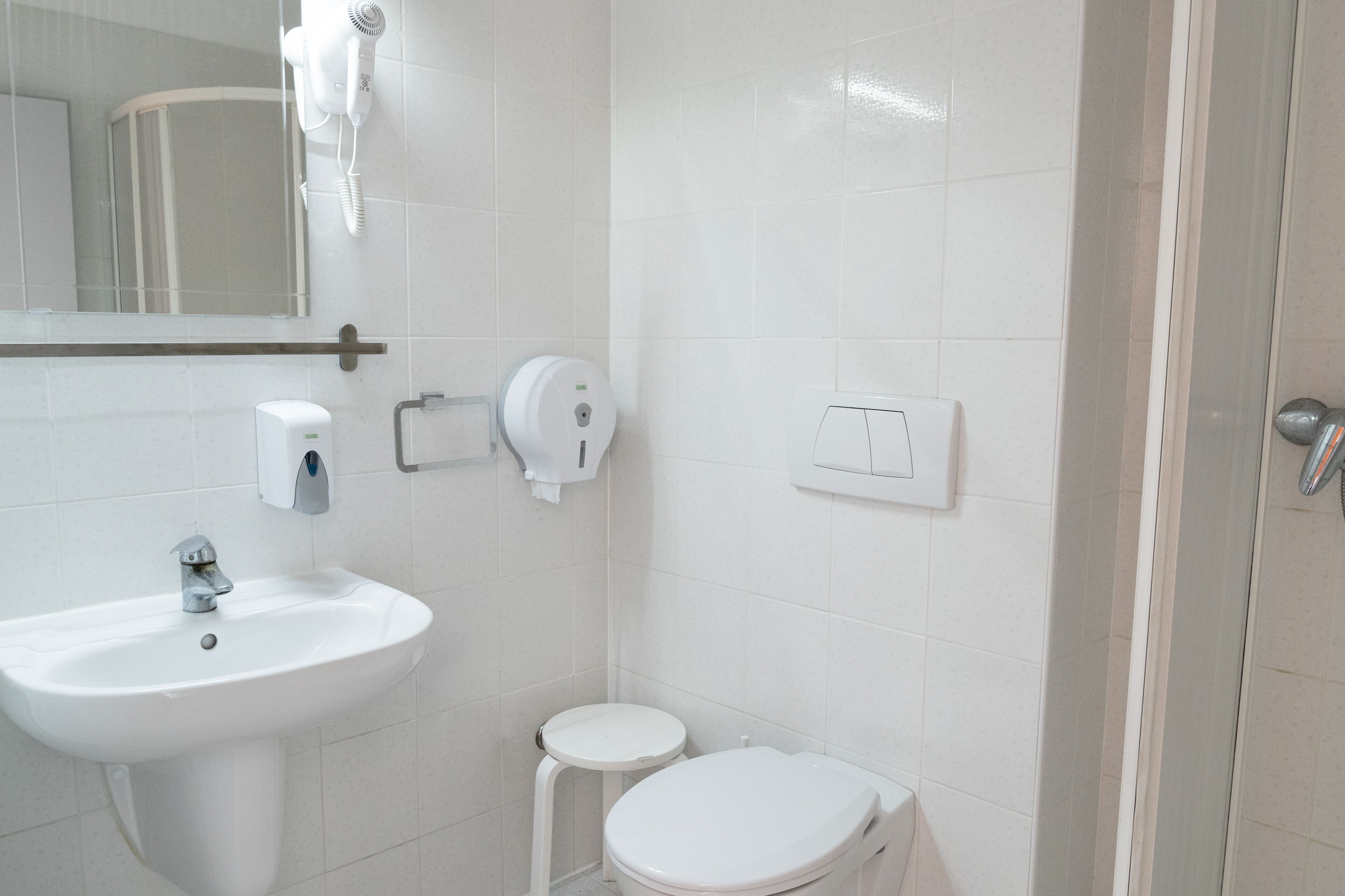 A bathroom with a white sink, toilet, and a mirror. There is a hairdryer mounted on the wall and a soap dispenser.