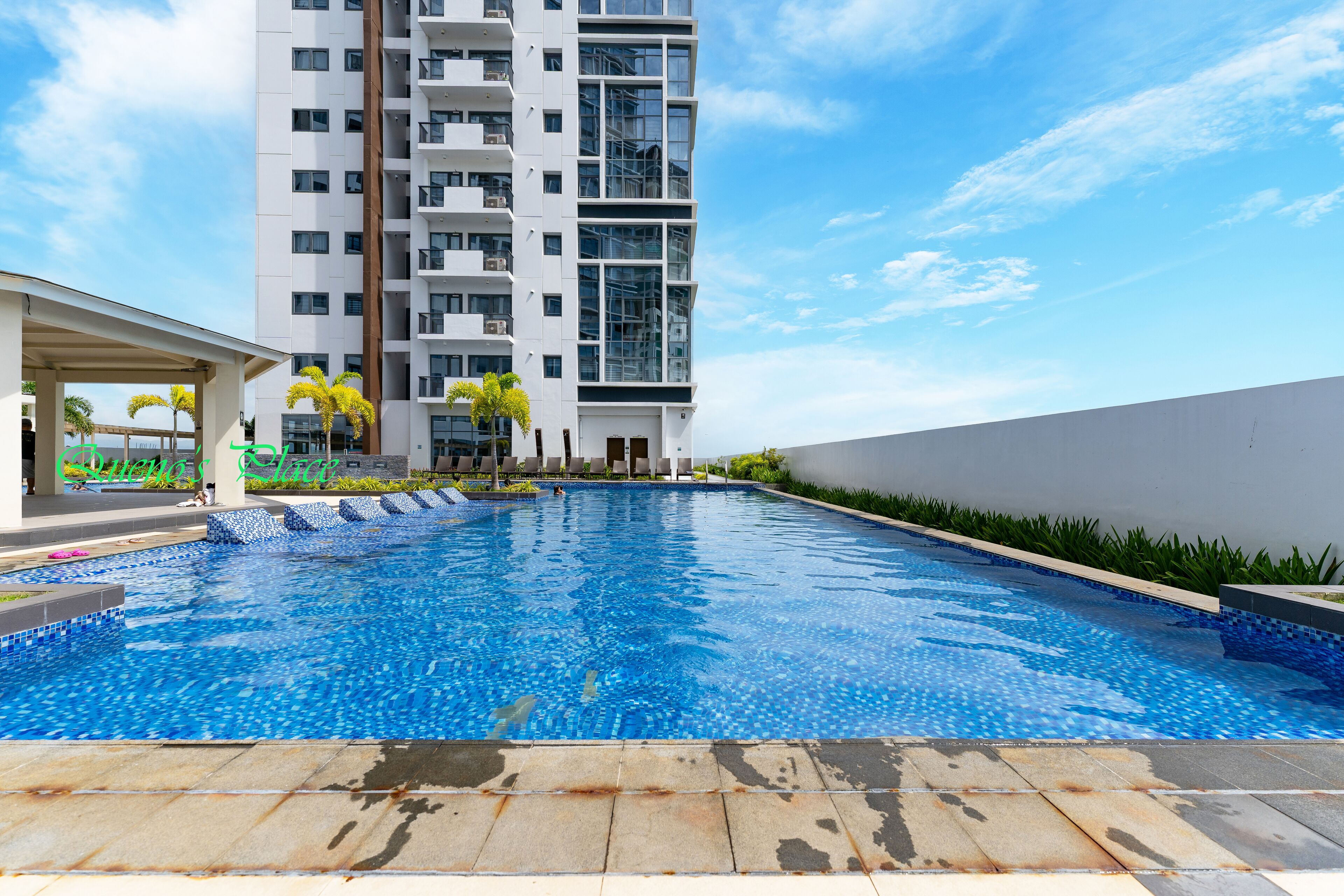 Una piscina con bordeado de baldosas, rodeada por un edificio de gran altura y una zona cubierta con palmeras.