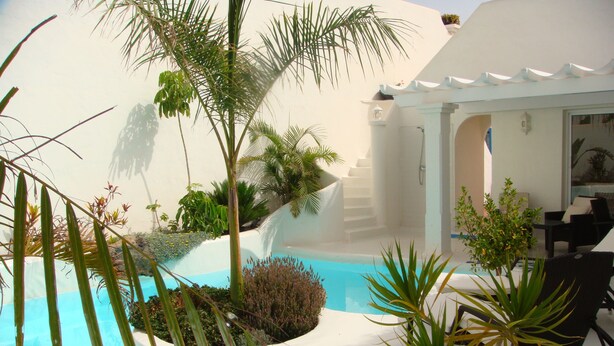 Una piscina rodeada de plantas tropicales y un edificio blanco con zona de estar techada.