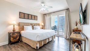 Une chambre d’hôtel avec un grand lit, deux lampes de chevet, un ventilateur de plafond, un téléviseur à écran plat et un balcon donnant sur un immeuble.