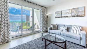 Un salon avec une table basse en verre, un canapé gris agrémenté de coussins à rayures et un balcon offrant une vue sur la mer.