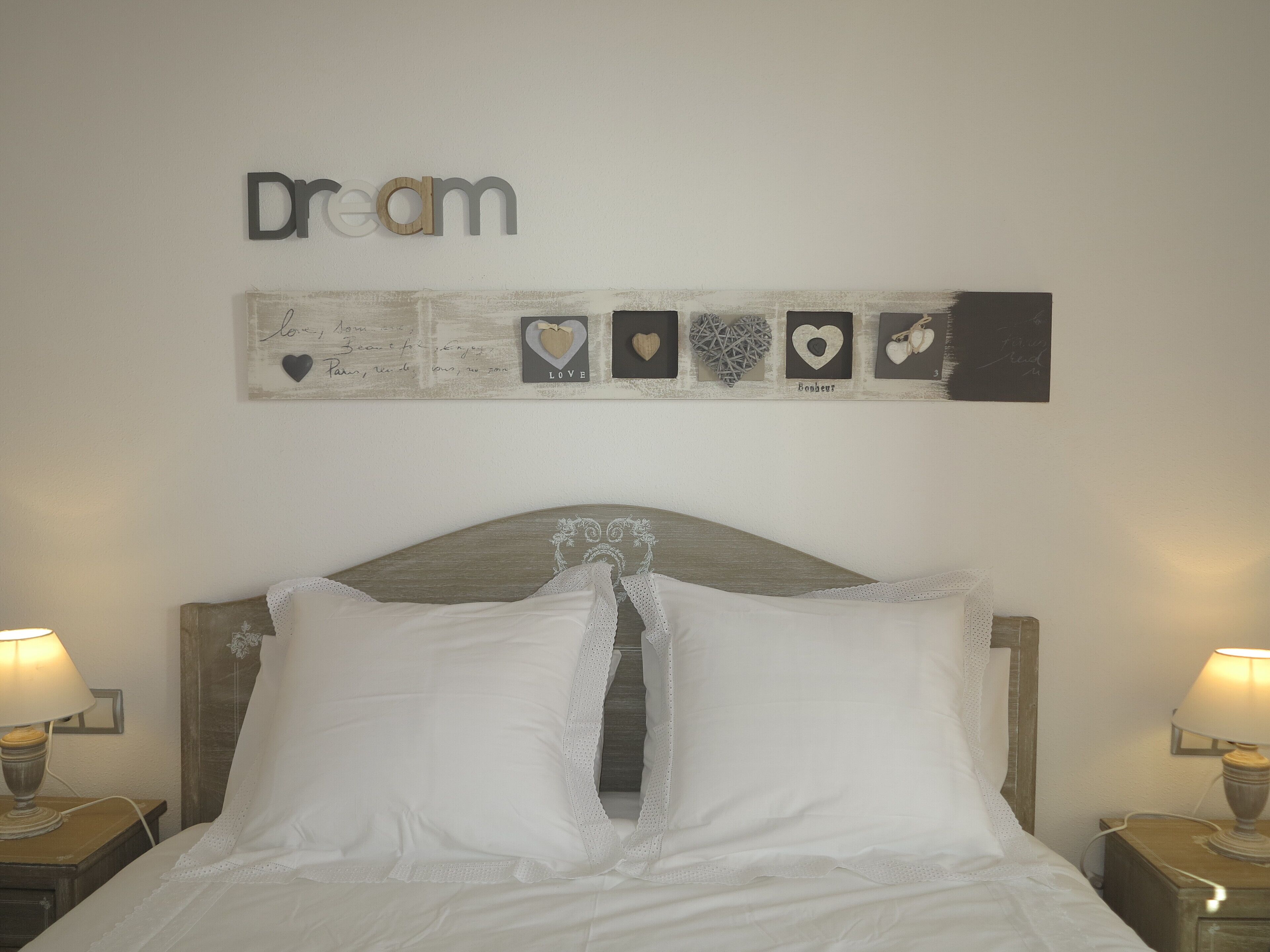 Ein ordentlich bezogenes Bett mit weißen Leinen und Spitzenakzenten, ein hölzernes Kopfteil und eine Wand mit einem 'Dream'-Schild und herzförmigen Verzierungen.
