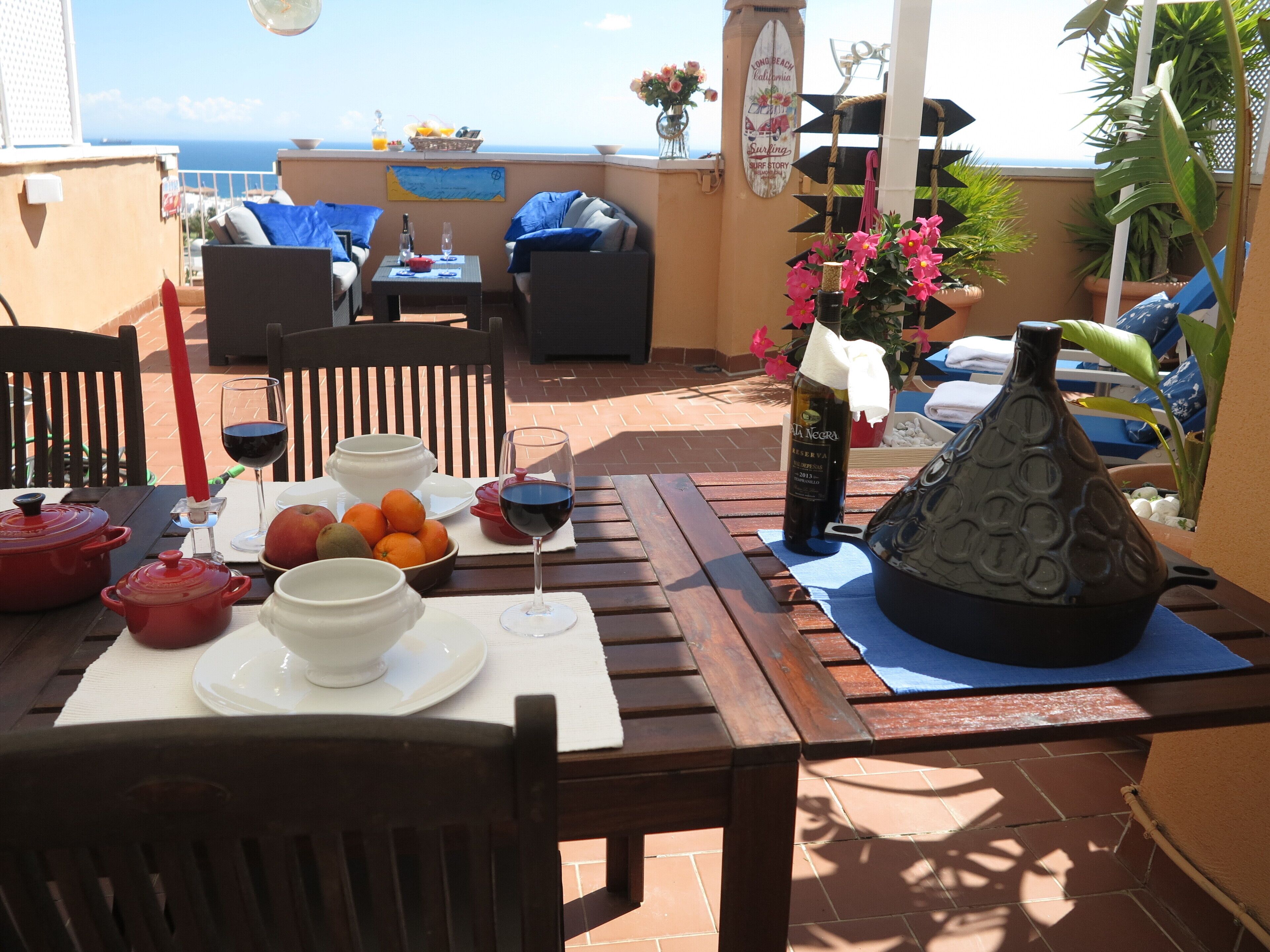 An outdoor dining area with a table set for two, featuring a bottle of wine, glasses, bowls, and fruits.